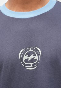 Εγγύς πλάνο ενός ατόμου που φορά μοβ πουκάμισο με γαλάζιο γιακά και λευκό αφηρημένο λογότυπο κύματος και κύκλου στο στήθος.