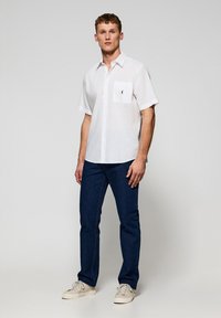 Camicia bianca a maniche corte con bottoni e taschino, abbinata a jeans blu scuro e sneakers color crema, in piedi su uno sfondo neutro.
