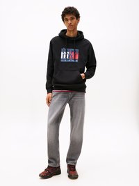 Felpa nera con logo "Tommy Jeans" in rosso, bianco e blu; abbinata a jeans grigi e scarpe nere con dettagli rossi.
