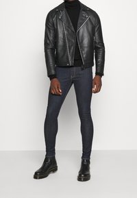 Veste de moto en cuir noir avec des fermetures éclair argentées, col roulé noir, jean slim bleu foncé et bottines noires avec des accents jaunes.