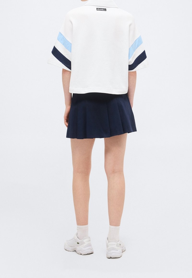 Personne debout de dos, portant un haut court blanc à rayures bleues, une jupe plissée bleu marine, des chaussettes blanches et des baskets blanches.