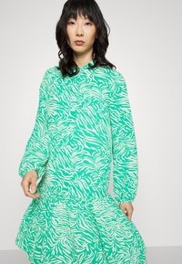 Vestido verde com riscas de zebra brancas, mangas longas, frente com botões e um acabamento franzido na barra. Feito de um tecido leve com uma textura suave.