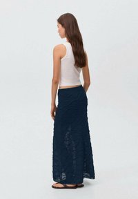 Jupe maxi tricotée bleu marine avec un motif en vagues, portée sur un top sans manches blanc. Caractéristiques d'une taille extensible et de fentes latérales. Sandales plates.