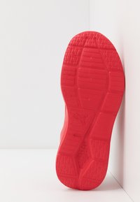 Semelle de chaussure de sport rouge présentant des motifs de grip texturés et un logo central, avec un profil plat et lisse et une partie de la pointe arrondie.