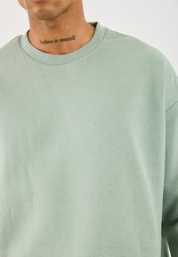 Hombre con una camiseta de cuello redondo de color verde claro y un tatuaje que dice "cree en ti mismo" en la parte frontal de su cuello, contra un fondo liso.