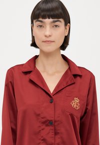 Bordo satīna krekls ar pogām un apkakli, ar kreisajā krūtī esošu kabatu un izšūtu zelta kroņa logotipu. Gluda tekstūra un brīva piegriezuma.