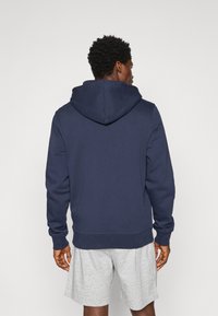 Marineblaues Hoodie mit großer Kapuze und gerippten Bündchen, aus einem weichen Material gefertigt. Getragen mit grauen Shorts, von hinten betrachtet.