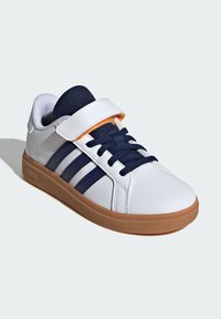 Zapatillas blancas con acentos en azul marino, que presentan una amplia correa de Velcro, una lengüeta de tejido texturizado y una suela de goma color caramelo. Diseño de tres rayas.