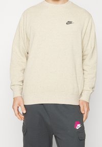 Bežová mikina s texturovaným povrchem. Na hrudi se nachází černé logo Nike. Manžety a spodní lem jsou žebrované pro těsné usazení. V kombinaci s tmavě šedými kalhotami.