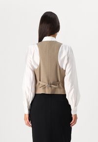 Tan vest med bagbånd over en hvid bluse med pufærmer. Tøjet er lagdelt over sorte bukser.