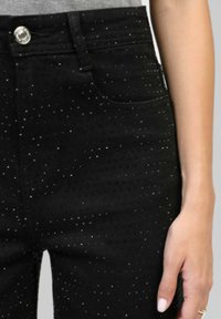 Gros plan sur un pantalon noir orné de petits clous argentés porté par une personne avec une bague en argent sur sa main reposant à côté de la cuisse.