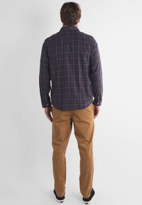 Homme de dos portant une chemise à manches longues à carreaux gris foncé, un pantalon beige et des chaussures noires, sur un fond blanc uni.