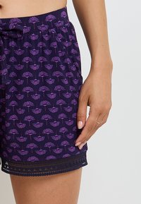 Shorts violets avec un motif floral en violet clair, dotés d'une taille à cordon et d'une finition en dentelle le long de l'ourlet. Tissu doux et léger.