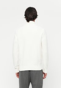 White knitted sweater with ribbed collar and cuffs, featuring a relaxed fit. The fabric has a soft, textured appearance.