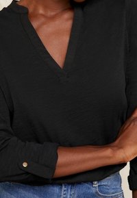 Musta, pitkähihaista pusero, jossa on V-pääkohdan muotoilu. Sisältää tekstuuripinnoitteen ja nappikiinnityksellä varustetun hihansuun. Rentoa mallia.