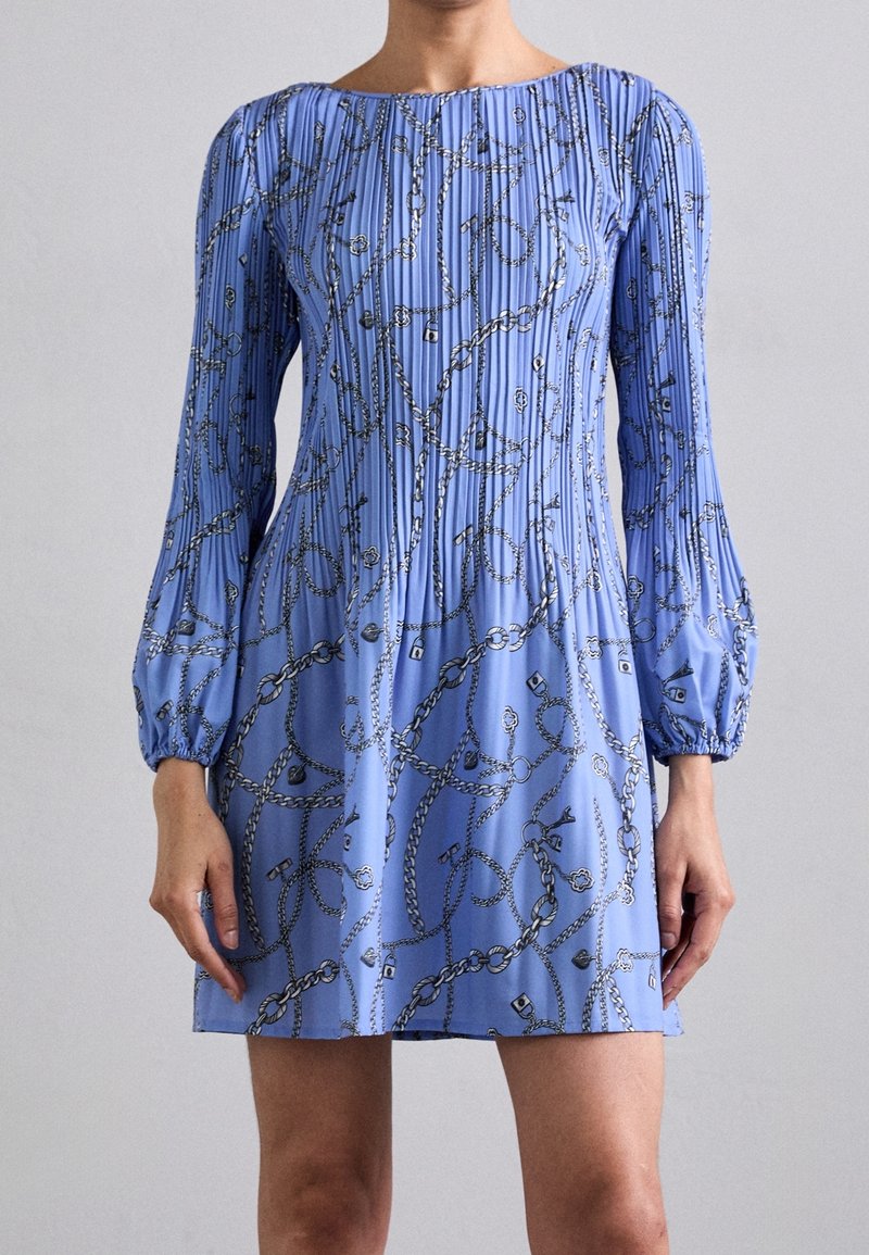 Femme portant une mini-robe bleue à manches longues avec un motif chaîne et des plis verticaux, debout devant un fond gris clair uni.