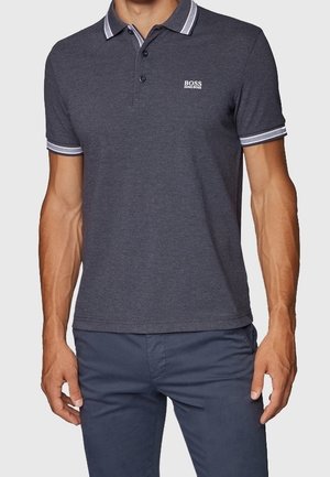 Mann trägt ein dunkelgraues Hugo Boss Polohemd mit weißer Paspelierung am Kragen und an den Ärmeln, kombiniert mit einer dunkelblauen Hose.