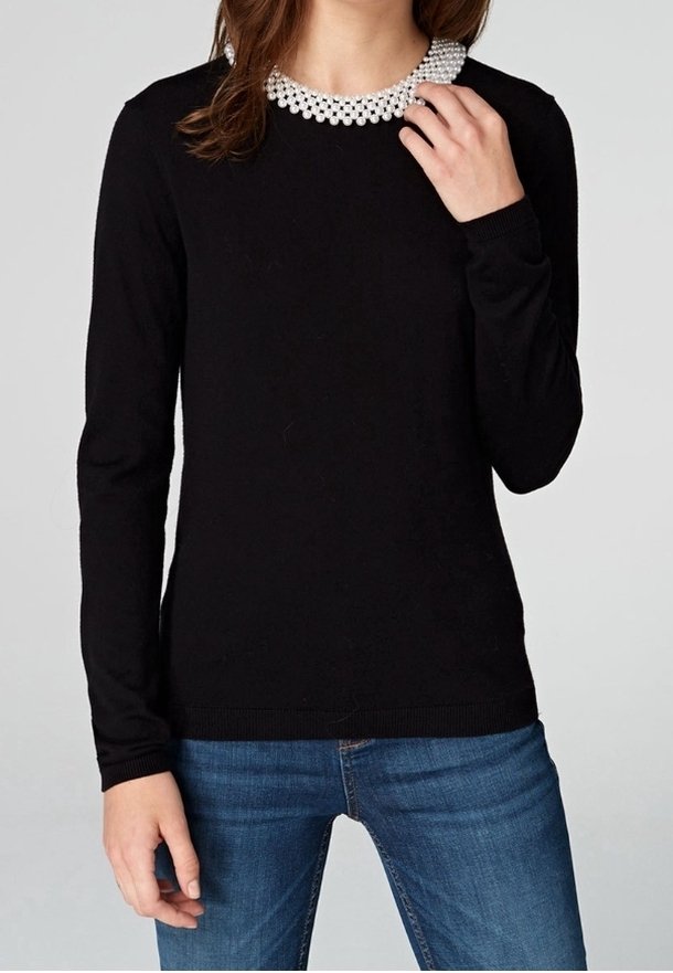 Svart långärmad tröja med rund halsringning, med detalj av vit pärlkrage. Tyget verkar mjukt och åtsittande. Bärs med blå jeans.