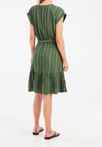 Grün gemustertes Kleid mit kurzen Ärmeln, Gürteldetail und einem gestuften Rüschensaum. Hat vertikale Streifen und kleine weiße Akzente.