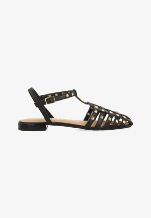 Sandalia de cuero negra con un diseño abierto, adornada con tachuelas doradas en forma de pirámide y una correa ajustable en el tobillo. Tacón plano y forma de punta.
