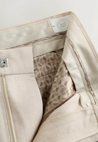Détail de la ceinture d'un pantalon beige montrant une fermeture éclair, une agrafe, un bouton intérieur et une doublure en tissu à motifs.