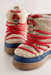 Stivali in pile beige da inverno con lacci rosso acceso, puntali blu e suole di gomma marrone visti dall'angolo frontale.