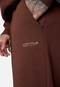 Brauner, gerippter Sweatshirt mit einer Seitentasche. Verfügt über das silberne "ESTEEM®"-Logo, das an der unteren Vorderseite gestickt ist. Glatte Textur, körperbetonter Schnitt.