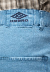 Κοντινό πλάνο σε ανοιχτό μπλε τζιν στο πίσω μέρος της μέσης με μαύρο λογότυπο Umbro και μέρος από καφέ καρό πουκάμισο που έχει βγει μέσα.