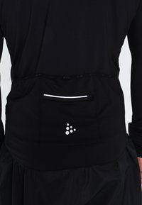 Černý sportovní top se malou zipovou kapsou, texturovaným vzorem a reflexním logem vzadu poblíž pásu.