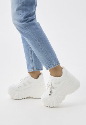 Persona con vaqueros azul claro y zapatillas blancas de plataforma con adornos decorativos plateados sobre un fondo blanco liso.