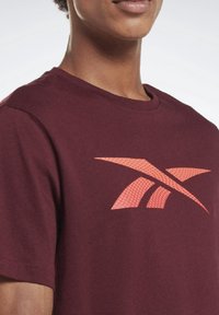 Camiseta de algodón color marrón con cuello redondo, que presenta un gran diseño de logo naranja con puntos en la parte frontal, textura suave y mangas cortas.