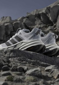 Λευκά παπούτσια τρεξίματος μονοπατιού Adidas Terrex με μαύρες ρίγες σε βραχώδες έδαφος κάτω από καθαρό ουρανό, αναδεικνύοντας τον ανθεκτικό σχεδιασμό και τις σόλες με κράτημα.
