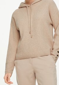 Sweat à capuche en tricot côtelé beige avec cordon de serrage et embouts dorés ; manches longues avec boutons décoratifs aux poignets.