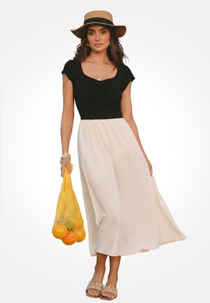 Donna che indossa un vestito bianco e nero con un cappello di paglia, che tiene una borsa gialla a rete con arance, in piedi con sandali beige.