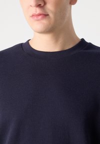 Tengerészkék textúrált pulóver, kerek nyakkal és bordázott anyaggal, a mellkastól felfelé látható. Nincsenek látható további minták vagy hardverek.