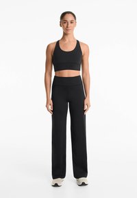 Schwarzes sportliches Bustier mit gerafftem Bund, kombiniert mit hoch taillierten, weiten schwarzen Hosen und hellen Sneakers. Glatte Textur.