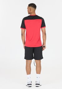 Punainen ja musta urheiluasu, lyhythihainen paita mustalla yoke-tyylillä, yhdistetty mustiin shortseihin. Sisältää näkyvän logon ja ranteeseen kiinnitettäviä asusteita.