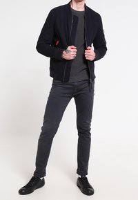 Giacca bomber marina con zip, polsini a costine e un accento rosso. Indossata sopra un maglione grigio testurizzato e jeans grigio scuro, abbinata a scarpe nere.