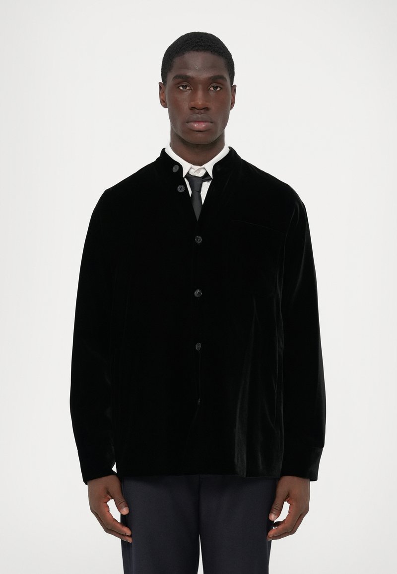 Black velvet jacket with a high collar, front buttons, and a chest pocket. Paired with a white shirt and tie, displaying smooth texture.