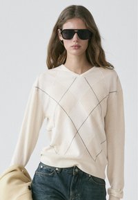 Maglione color crema a scollo a V con motivo a diamante delineato in nero; maniche lunghe; tessuto morbido. Indossato con jeans di denim scuro e un cappotto beige.