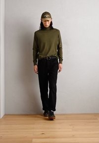 Maglietta verde oliva a maniche lunghe con colletto a dolcevita abbinata a pantaloni neri e un cappello coordinato; le calzature hanno dettagli verde scuro e una suola marrone.