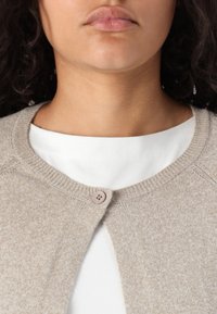 Beige strikket cardigan med rund hals, med en enkelt knappelukking og fine ribbingsdetaljer langs kanten. Brukt over en hvit skjorte.