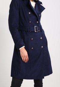 Mörkblå dubbelknäppt kappa med bälte i midjan, silverfärgade knappar och slagkrage. Materialet verkar mjukt och texturerat.