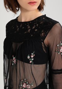 Blusa nera trasparente con dettagli in pizzo sul collo e ricami floreali in rosa e verde sulle maniche. Tessuto texturizzato con un design fluido.