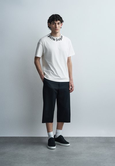 Νεαρός άνδρας στέκεται μπροστά σε απλό ανοιχτό γκρι τοίχο, φορώντας λευκό μπλουζάκι με γραφικό σχέδιο, μαύρο φαρδύ παντελόνι έως τον αστράγαλο, λευκές κάλτσες και μαύρα αθλητικά παπούτσια.