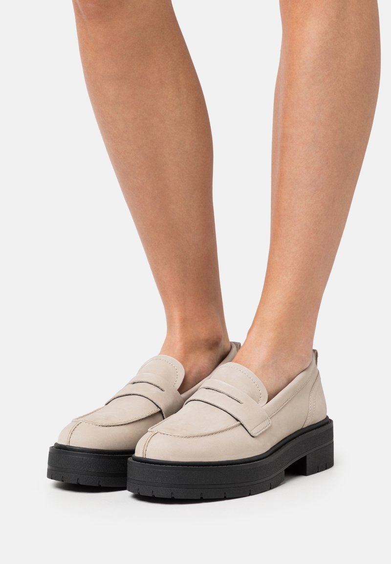 Mocassins en cuir beige avec une semelle noire épaisse, présentant un détail de sangle cousu sur le devant et une texture lisse.