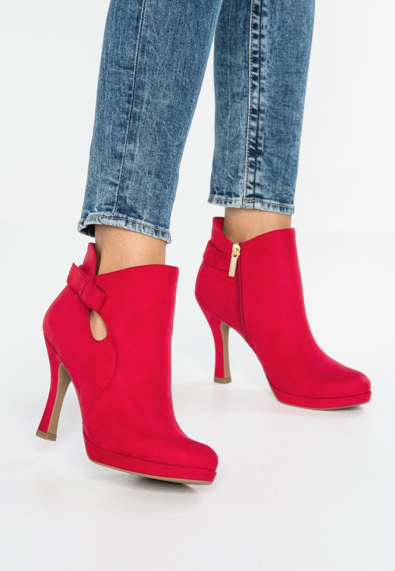 Jambes en jean bleu portant des bottines à talons hauts rouges vives avec fermeture éclair latérale et petite découpe près de la cheville.