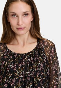 Musta kukkainen paita, jossa on rypistetty pääntie, läpinäkyvä kangas, jossa on pieniä vaaleanpunaisia ja vihreitä kukkakuoseja kauttaaltaan.