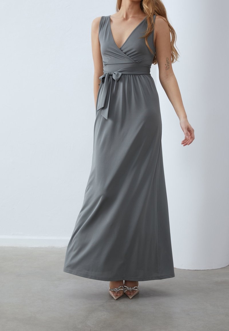 Pitkä, kääntötyylinen mekko pehmeästä harmaasta kankaasta, syvällä V-pääntiellä, istuvalla vyötäröllä ja solmittavalla vyöllä. Sileä pinta ja virtaava siluetti.