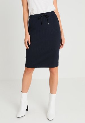 Femme portant une jupe côtelée bleu marine longueur genou avec taille à cordon, un top blanc sans manches et des bottines blanches à talons carrés.